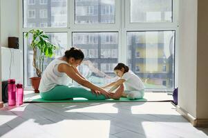 Jeune femme exercice avec sa fille, élongation sa corps contre le Contexte de grand les fenêtres à Accueil sur une magnifique ensoleillé journée photo