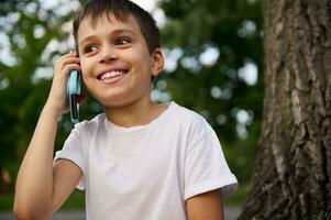 de bonne humeur adorable école enfant garçon parlant sur mobile téléphone, mignonne souriant à la recherche loin, repos sur le Publique parc après premier journée à école sur magnifique été journée. concepts de retour à école photo