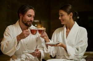 content nouveau marié, charmant milieu vieilli marié caucasien couple dans blanc peignoirs en portant tasses avec à base de plantes thé, séance proche à chaque autre, visage à affronter, profiter temps ensemble dans bien-être spa recours photo