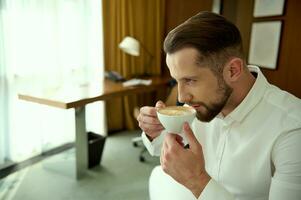 sur de soi attrayant Beau Jeune homme d'affaire les boissons fraîchement brassée café pour bien début de le jour, séance dans Hôtel pièce pendant le sien affaires voyage photo