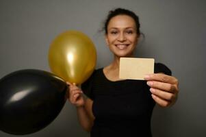 doux concentrer occus sur vide Vide d'or crédit carte dans le main de magnifique souriant femme en portant noir or air des ballons, posant contre gris mur Contexte avec copie un d espace. noir Vendredi concept photo