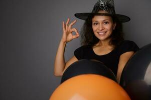 fermer de magnifique souriant hispanique femme dans sorcier chapeau, habillé dans sorcière carnaval costume pour Halloween faire la fête, spectacles D'accord signe, pose contre gris Contexte avec noir des ballons, copie espace photo