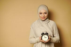 mignonne Jeune musulman femme dans hijab en portant alarme l'horloge dans mains et souriant mignonne à caméra permanent contre une beige Contexte avec copie espace. photo