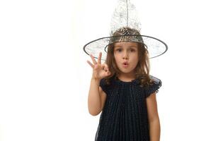 isolé taille longueur portrait de jolie peu sorcière fille portant une sorcier chapeau et habillé dans élégant carnaval robe, faire des gestes, montrant D'accord signe avec des doigts. Halloween concept sur blanc Contexte photo