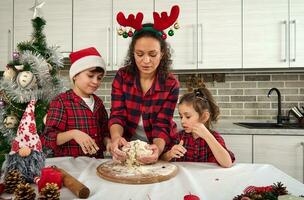 peu des gamins portion leur maman à pétrir une pâte sur une en bois conseil, cuisine à Accueil cuisine pour Noël traditionnel événements. magnifique mère enseignement sa fils et fille Comment à préparer le pâte photo