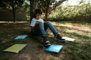 hispanique adorable diligent adolescent écolier dans décontractée denim vêtements, séance en dessous de le arbre dans une parc et en train de lire livre photo