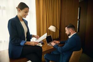 affaires les partenaires travail ensemble dans Hôtel pièce sur affaires voyage. femme d'affaires, patron avec empiler de papier les documents sourires, en train de lire contact, Beau collègue séance à table et travail sur portable photo