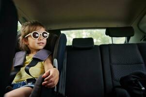 une magnifique fille dans des lunettes de soleil est assis dans une enfant voiture siège dans le voiture et regards en dehors le la fenêtre. sûr Voyage avec les enfants dans le voiture photo