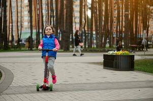 plein longueur portrait de un européen mignonne enfant fille équitation une donner un coup scooter dans le parc sur une ensoleillé journée dans de bonne heure l'automne ou printemps. content enfance concept photo