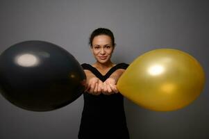 magnifique femme habillé dans noir sourit à la recherche à caméra et pose contre gris Contexte en portant or et noir air des ballons dans sa tendu main. noir Vendredi concept avec copie un d espace photo