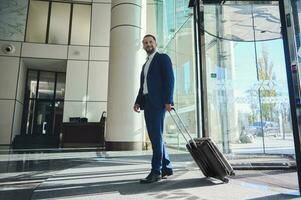 une toute la longueur Beau homme d'affaire développeur entrepreneur dans une affaires costume avec une valise des stands à le entrée de une moderne luxe bâtiment, en marchant à le hall pendant le sien affaires voyage photo