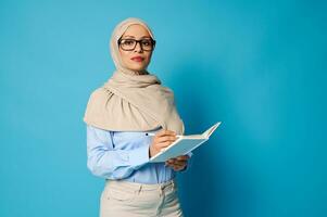 sérieux femme dans hijab et des lunettes avec stylo et livre posant sur bleu Contexte avec copie espace photo
