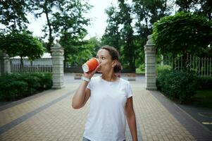 charmant Jeune femme en buvant à emporter café de une jetable papier carton tasse, tandis que profiter sa marcher dans parc photo