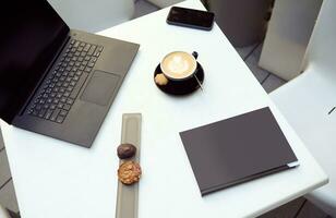 plat allonger composition de délicieux des pâtisseries sur une blanc table suivant à une portable et une tasse de délicieux aromatique cappuccino café, une mobile téléphone et une noir relié livre. copie un d espace photo