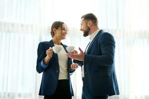 charmant femme et Beau homme dans marine affaires costume ayant une conversation plus de une tasse de café pendant une travail casser, discuter des plans et buts, permanent par le fenêtre et souriant à chaque autre photo