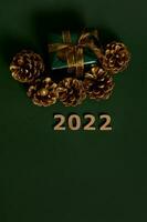 Haut vue de Noël présent dans briller emballage papier avec d'or arc et pins cônes coloré dans or, chiffres dans bois symbolisant le année 2022 posé en dehors sur foncé vert Contexte avec copie espace photo