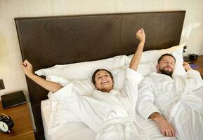 content marié couple, jeunes mariés, magnifique épouse et aimant mari, les amoureux élongation dans le chambre après veille en haut, émouvant chaque autres mains et souriant, profiter le intimité sur leur voyage de noces photo