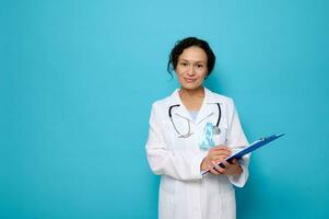 sur de soi portrait de milieu vieilli femelle mixte course médecin dans blanc médical robe, portant une bleu conscience ruban, pose avec une presse-papiers dans sa mains, à la recherche à appareil photo, copie espace pour médical un d photo