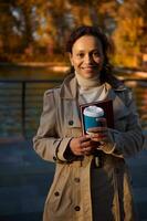 portrait de une de bonne humeur africain femme avec à emporter tasse de café et une main livre souriant à la recherche à caméra sur le l'automne parc Lac Contexte avec chute feuilles. automnal Extérieur loisirs. des loisirs photo