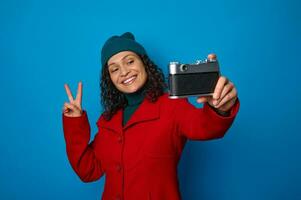 content photographe, attrayant souriant africain femme dans rouge manteau et vert chapeau montrant paix signe tandis que prise une autoportrait avec une ancien caméra, isolé sur bleu Contexte avec copie un d espace photo