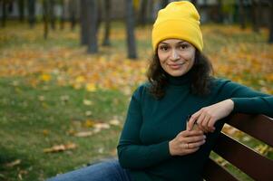 confiant Enchanté femme dans brillant coloré chaud confortable vêtements repos sur une parc banc avec une papier carton café tasse sur le Contexte de automnal érable et chêne des arbres, mignonne sourit à la recherche à caméra photo