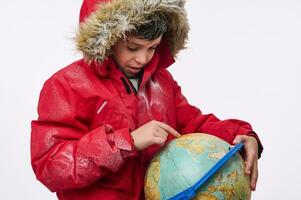 adorable élémentaire vieilli caucasien garçon dans une chaud couvert de neige rouge vers le bas veste avec une capot, montrer du doigt le sien doigt à le globe dans chercher de une Voyage destination. hiver tourisme concept avec copie un d espace photo