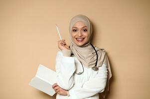 arabe musulman femelle médecin en portant une journal intime et à la recherche à caméra avec une magnifique à pleines dents sourire , montrer du doigt avec une blanc crayon sur une beige Contexte avec copie espace. photo