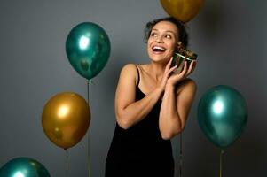 attrayant femme dans soir robe détient une Noël cadeau près sa affronter, doucement câlins il, regards en haut sur copie espace sur gris Contexte avec or vert air des ballons. Nouveau année, anniversaire fête concepts pour un d photo