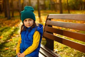 mode de vie portrait de jolie bébé fille dans coloré brillant chaud vêtements repos sur une parc banc, profiter Extérieur loisir et l'automne temps dans l'automne forêt parc à magnifique le coucher du soleil. en bonne santé mode de vie photo