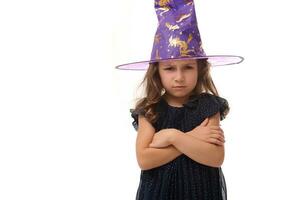 portrait de jolie peu sorcière en colère dérangé fille portant une sorcier chapeau et habillé dans élégant carnaval robe, à la recherche à caméra posant avec franchi bras contre blanc arrière-plan, copie espace. Halloween photo