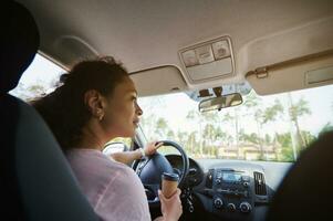 mature chauffeur femme à la recherche à le côtés et en mettant sa main avec à emporter papier tasse sur une pilotage roue dans le en mouvement voiture photo