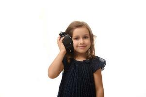 une magnifique 4 ans vieux peu fille dans une foncé bleu robe détient un alarme l'horloge près sa oreille et écoute attentivement à le son, sourit à la recherche à caméra, isolé sur blanc Contexte avec copie espace photo