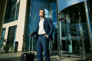 portrait de une réussi Jeune homme d'affaire portant décontractée affaires costume des stands avec une noir valise à le verre entrée à le hôtel, regards en toute confiance à le côté, en portant le sien main dans le sien poche photo