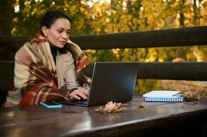 pigiste milieu vieilli femme, développeur enveloppé dans chaud de laine couverture, les types sur portable clavier, travaux projet, des plans Commencez, est assis à en bois table dans pays loger, travaux à distance sur cool l'automne journée photo