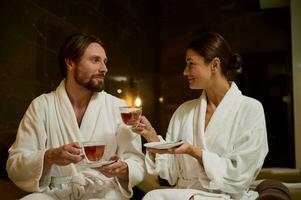 aimant couple dans blanc peignoirs séance proche à chaque autre sur une salon zone par le mer, en buvant en bonne santé à base de plantes thé avec de tasses. jeunes mariés sur leur voyage de noces profiter bien-être spa procédures photo