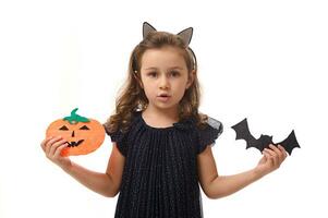 isolé portrait sur blanc Contexte avec copie espace de adorable peu fille, 4 ans vieux enfant, montrant à le caméra fait maison coupe-feutre chauve souris et citrouille dans sa mains, symbole de le Halloween fête photo