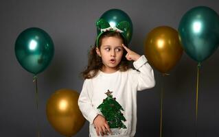 pensif fille met sa doigt sur sa temple et pensivement regards bas, posant contre gris Contexte avec magnifique d'or et brillant vert métallique air des ballons avec copie espace pour Noël un d photo