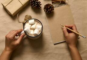 plat poser. Humain mains tenir une tasse avec chaud cacao boisson et guimauves et l'écriture avec en bois crayon sur Vide papier feuille de bloc-notes sur le papier carton surface avec artisanat Noël cadeau et pin cônes photo