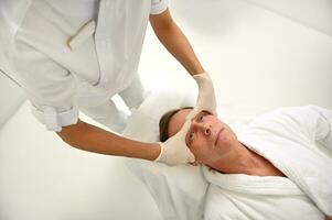 Haut vue de un méconnaissable esthéticienne performant une faciale levage lissage anti-âge professionnel massage de mature homme front. peau se soucier et Masculin beauté procédures concept à bien-être spa photo
