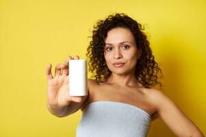 doux concentrer sur blanc récipient avec cosmétique produit dans main de magnifique moitié nu frisé femme enveloppé dans une baignoire serviette photo