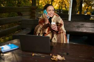 magnifique femme, développeur enveloppé dans une chaud plaid de laine couverture, pourparlers sur mobile téléphone, détient une à emporter papier tasse avec chaud café tandis que travail à distance sur ordinateur portable, Planification projets, début en haut photo