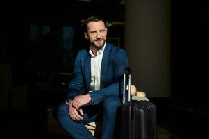 portrait de une Beau réussi homme, sur de soi homme d'affaire, souriant investisseur séance dans une chaise dans une Hôtel hall avec une bagage pendant affaires voyage photo