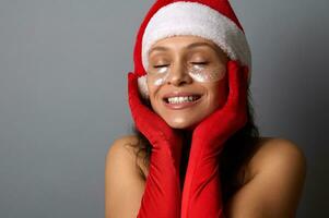 Jeune femme dans Père Noël carnaval tenue, avec brillant lissage taches en dessous de sa yeux, détient sa mains près sa affronter, sourit avec une magnifique à pleines dents sourire et pose avec fermé yeux sur une gris Contexte photo