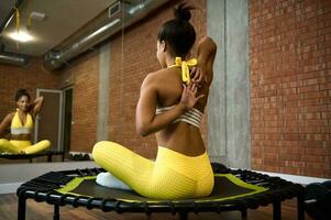 Jeune africain femme dans brillant Jaune survêtement élongation sa bras derrière sa dos, séance dans lotus position sur trampoline et à la recherche à sa miroir réflexion tandis que travail en dehors dans aptitude studio photo