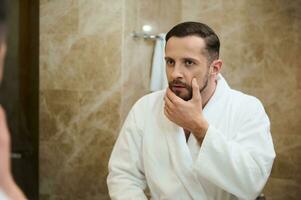 Beau Jeune européen homme dans une blanc éponge peignoir de bain émouvant le chaume avant rasage, à la recherche dans surprise à le sien miroir réflexion dans le salle de bains photo