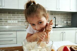 enfant prépare pâte, regards à le caméra penché contre une en bois cuillère pour pétrissage pâte. les enfants cuisine photo