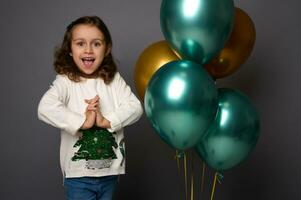 portrait sur gris Contexte avec copie espace pour Nouveau année La publicité de peu fille dans Noël blanc chandail, posant contre vert métallique et or air des balles, joliment souriant à la recherche à caméra photo