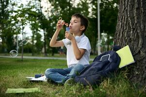 une écolier de élémentaire grades repos dans le parc séance sur le vert herbe après école, soufflant bulles. école sac avec classeurs et école Provisions mensonge vers le bas sur le herbe photo