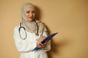 femelle musulman médecin portant beige hijab mignonne sourit à caméra et détient une stylo et presse-papiers dans mains. photo