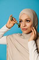 une Jeune magnifique musulman femme avec couvert diriger, Faire une faciale massage avec une jade rouleau. photo